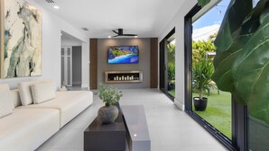 A modern living room with a sofa, a coffee table, a TV mounted on the wall, and a large window with a view of greenery. - 3 Bedroom Luxury Modern Home with Heated Pool and Near Downtown Dining & Shops! (Wilton Manors)