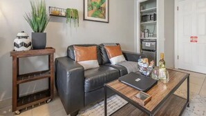 Een woonkamer met een zwarte leren bank, een houten salontafel, een laptop en een fles wijn. Er is een plank met planten en een inlijsting schilderij aan de muur.