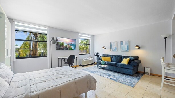 Ein Schlafzimmer mit einem Bett, einem Schreibtisch, einem Sessel, einer Couch und einem großen Fenster mit Blick auf Palmen.