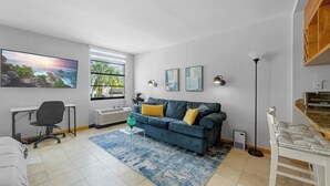 Ein Wohnzimmer mit einem blauen Sofa, einem Schreibtisch, einem Sessel, einem großen Fenster und Kunstwerken an der Wand.