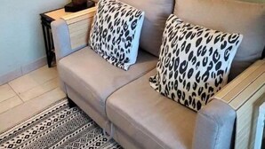 Un salón con sofá color beige, cojines de estampado de leopardo, alfombra estampada, un cuadro con jarrones y flores, y una escultura de madera.