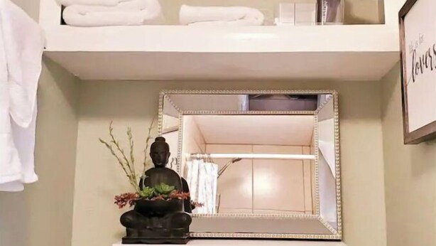 Un baño con repisas que sostienen toallas, un secador de pelo y artículos de tocador. Debajo de la repisa, hay un espejo con una estatua decorativa de Buda y una pequeña planta.
