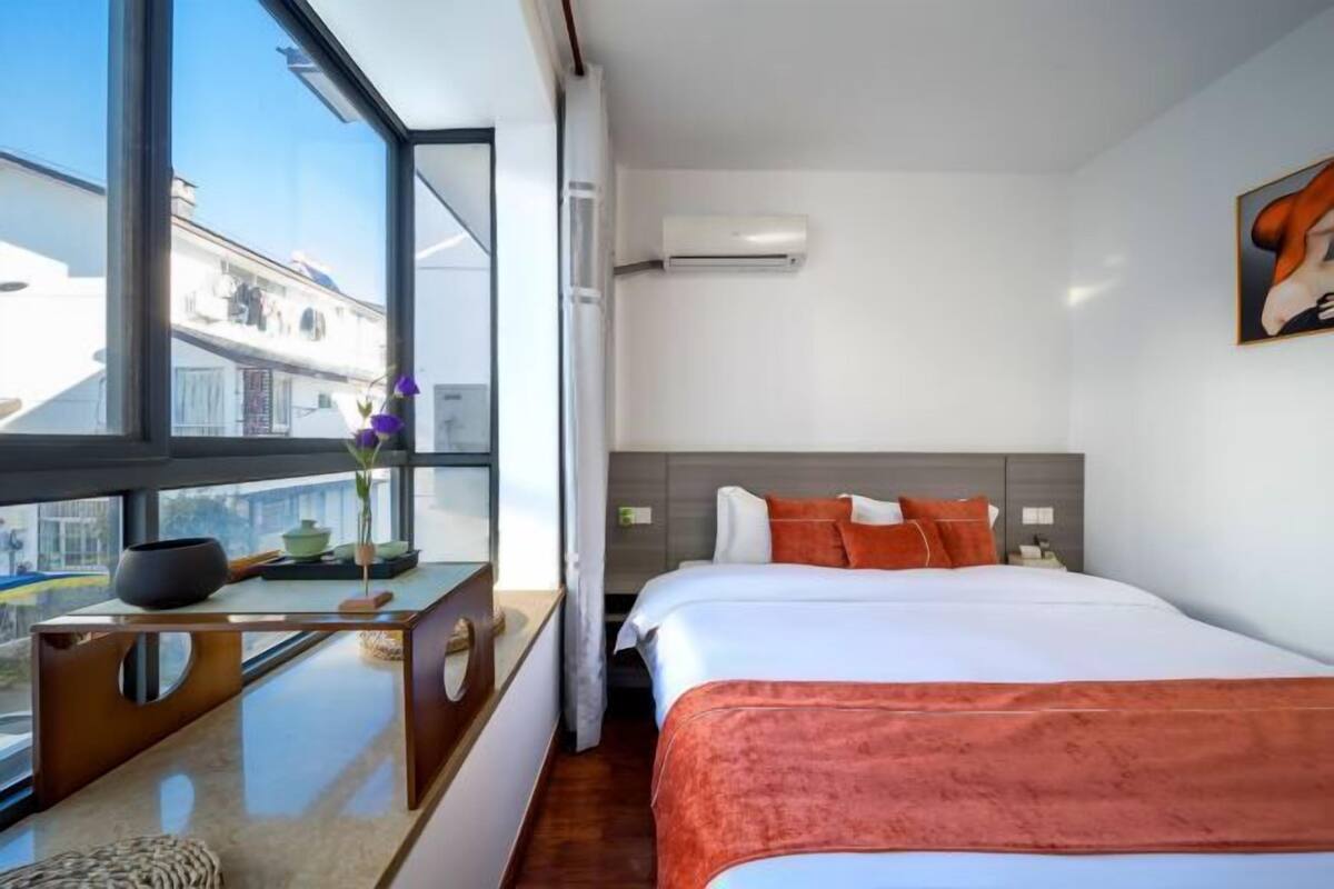 A modern hotel room with a large window, a bed with orange bedding, a bedside table with a vase of flowers, and a painting on the wall.
