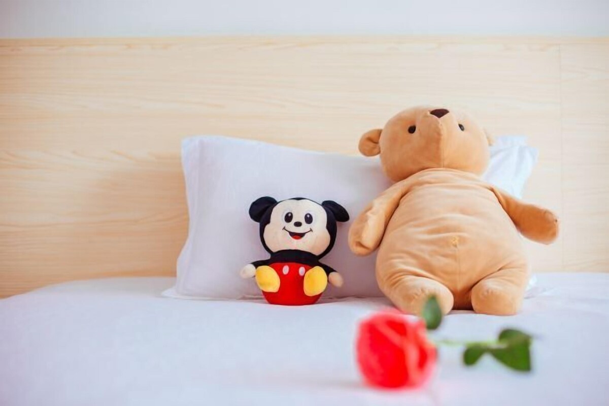 A bed with a white pillow featuring a cartoon character, a plush teddy bear, and a red rose.