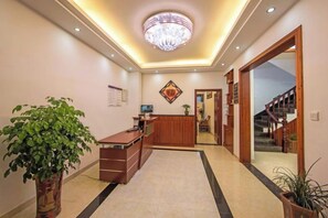 Zona de recepción de un hotel con mostrador, una planta en maceta, un candelabro y una escalera que lleva a los pisos superiores.