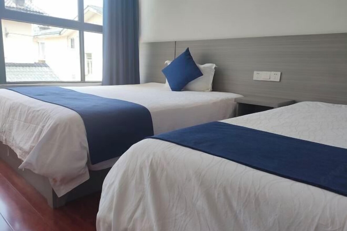A hotel room with two beds, each with a blue and white bedspread, and a window with a view of buildings outside.