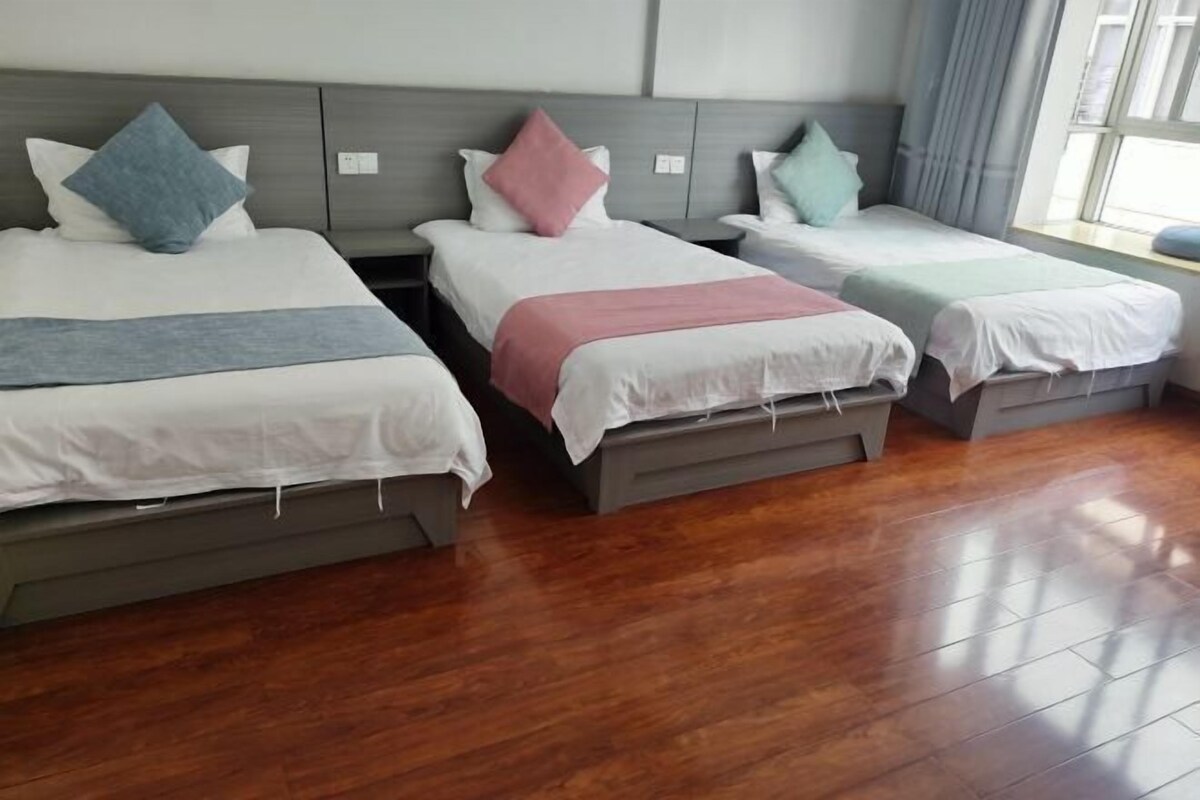 A hotel room with three single beds, each with a different colored pillow and blanket. The room has wooden flooring and a large window with curtains.