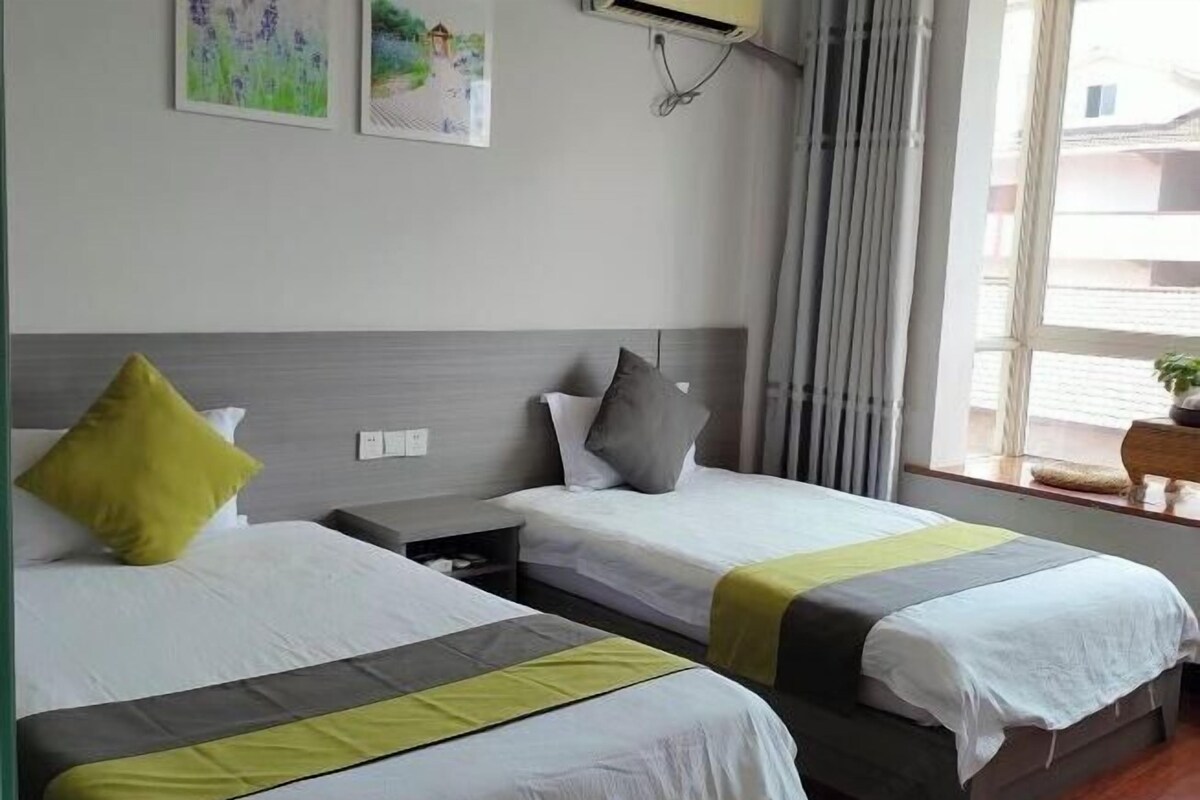 A hotel room with two beds, each with a grey and white bedspread, a yellow pillow on one bed, and a grey pillow on the other. There are two paintings on the wall, a window with curtains, and an air conditioning unit above the beds.