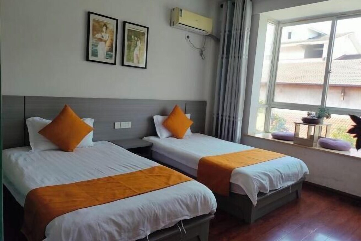 A hotel room with two beds, each with white bedding and orange accents, a window with a view of buildings, and an air conditioning unit on the wall.