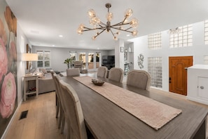 A dining area with a long table, chairs, a chandelier, and a view of the outside. - Luxury Beach Mansion - Roof Deck- Steps to Sand (Oxnard)
