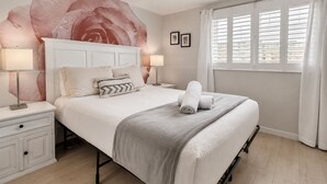 A hotel room with a large bed, two bedside tables with lamps, a large floral wall mural, a window with white curtains, and a framed picture on the wall. - Luxury Beach Mansion - Roof Deck- Steps to Sand (Oxnard)