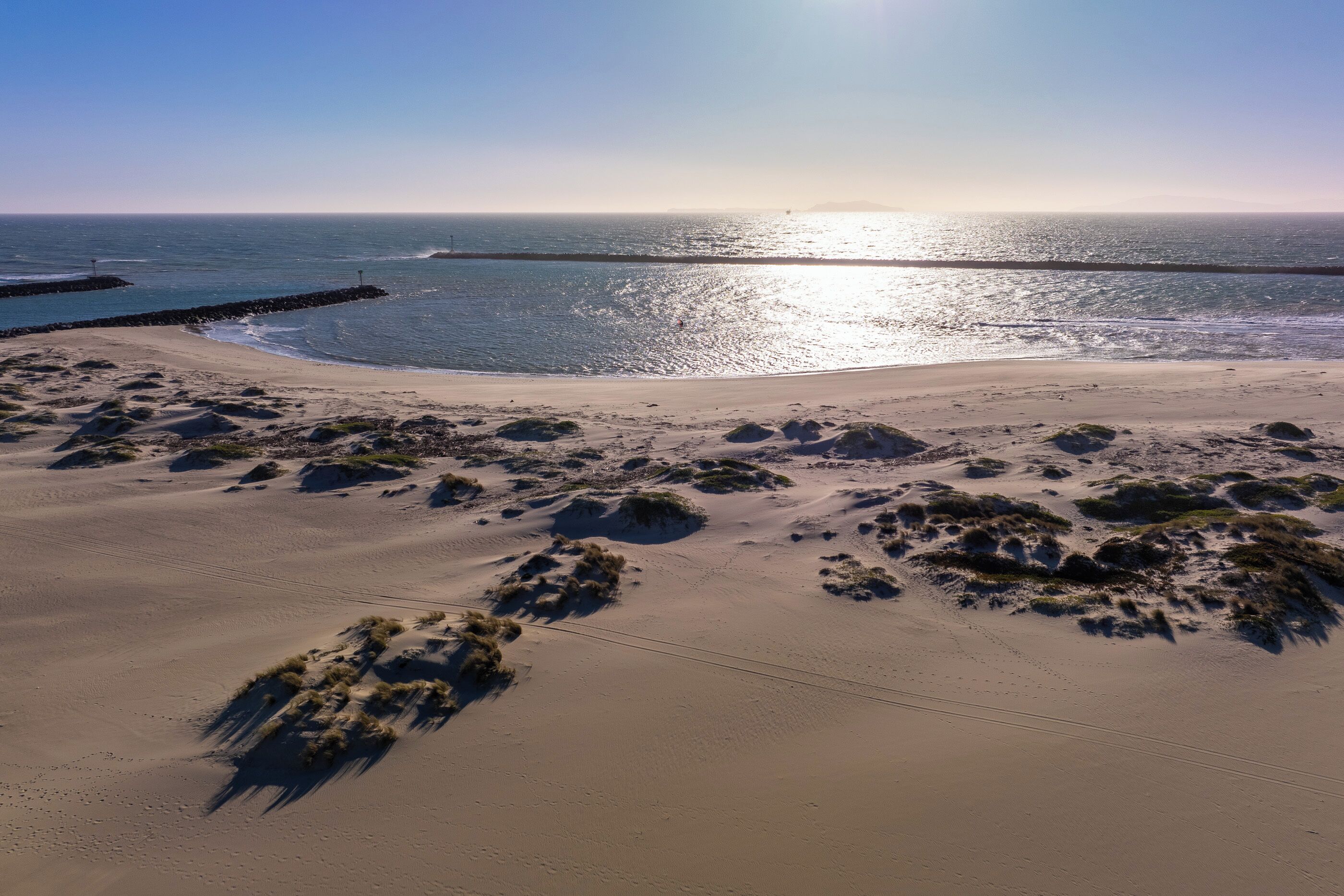 A coastal landscape with a sandy beach, sparse vegetation, and a clear sky.
