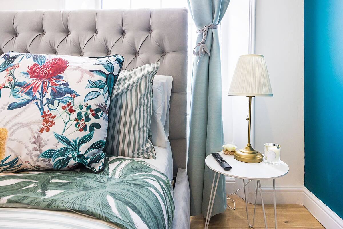 A neatly made bed with floral and striped pillows, a tufted headboard, a bedside table with a lamp, remote controls, and candles.