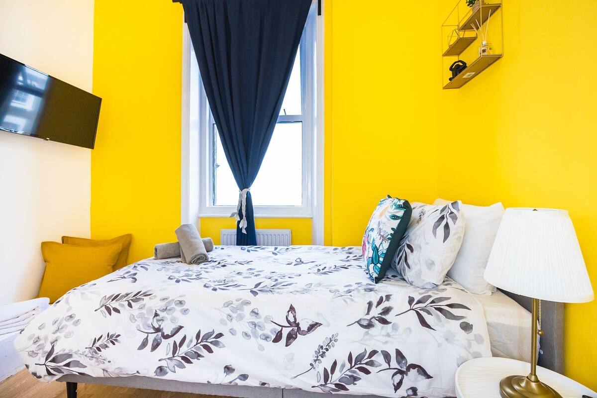 A bedroom with a bed, a bedside table, a lamp, a window with curtains, and a wall-mounted TV.