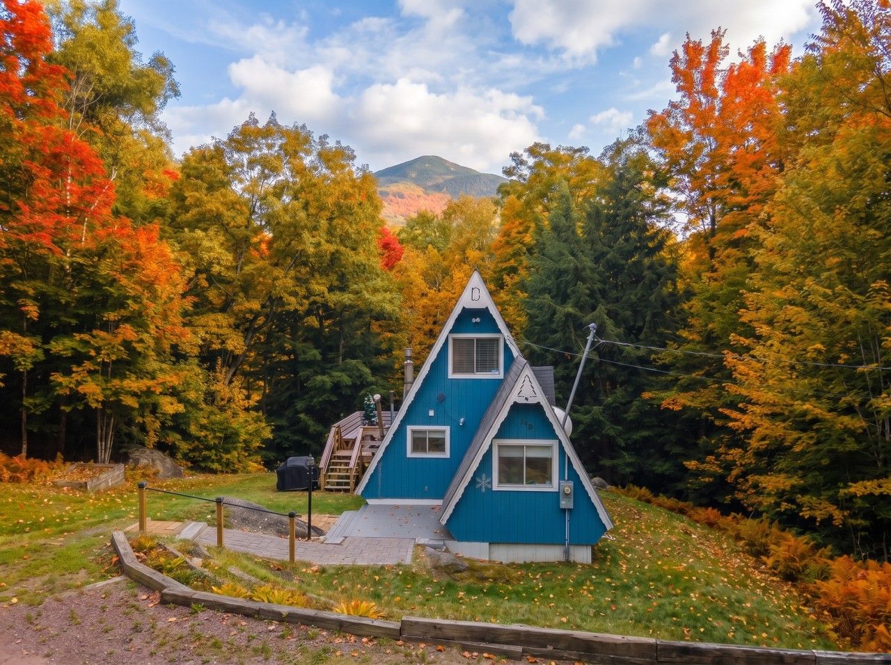A cabin with a triangular roof, surrounded by autumn foliage and a mountain in the background.