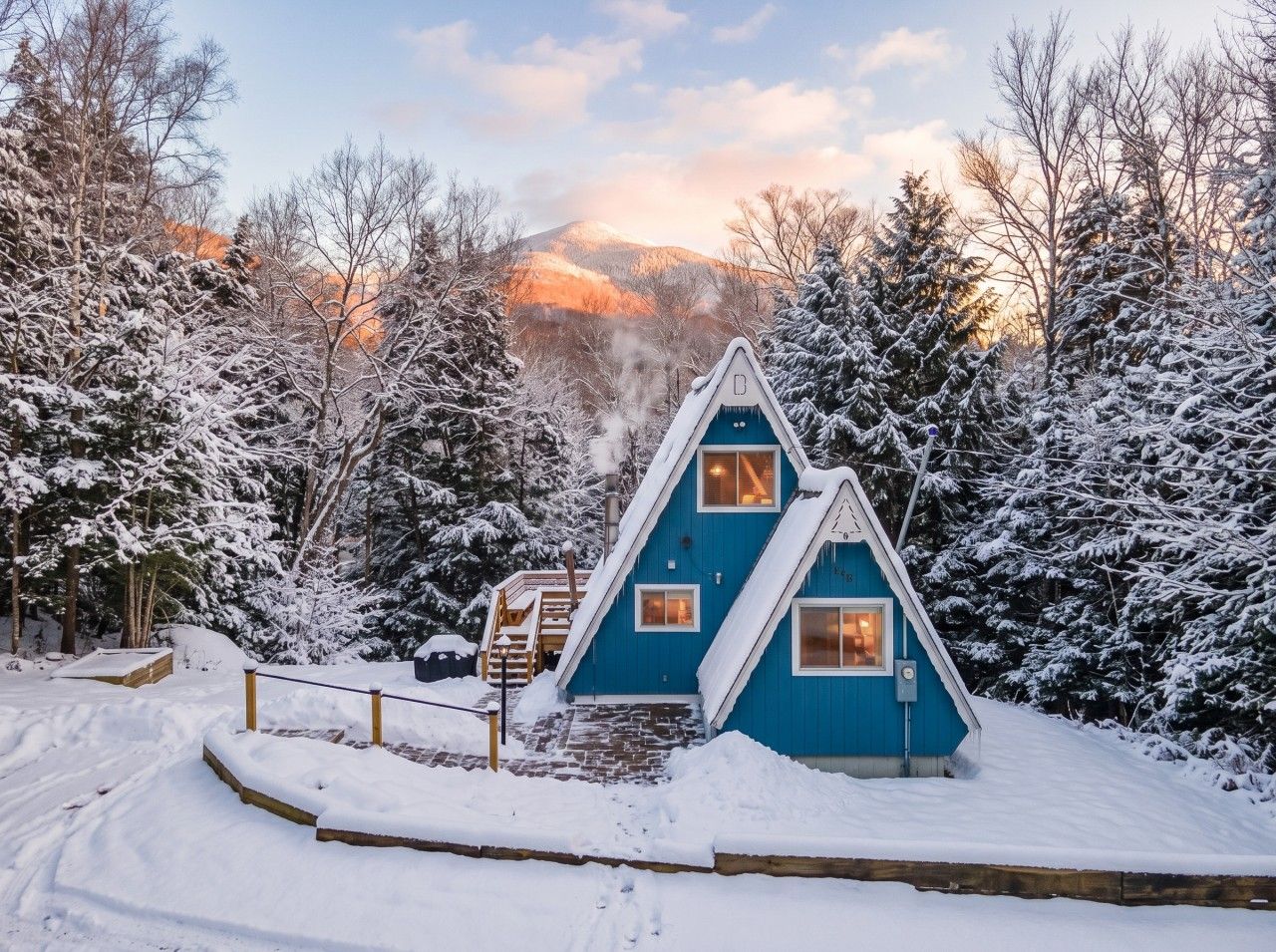 A snow-covered cabin with a steep roof, surrounded by trees and a mountain in the background.