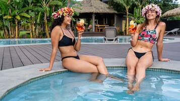 Two individuals in a pool with a tropical setting, including a thatched-roof structure and lush greenery in the background.