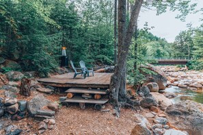 A rustic wooden deck with chairs, surrounded by rocks and trees, overlooking a river. - Stunning custom bld-riverside-pet/family friendly (Intervale)