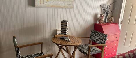 A room with striped wallpaper, a painting of birds on a fence, a red dresser, and a patterned chair.