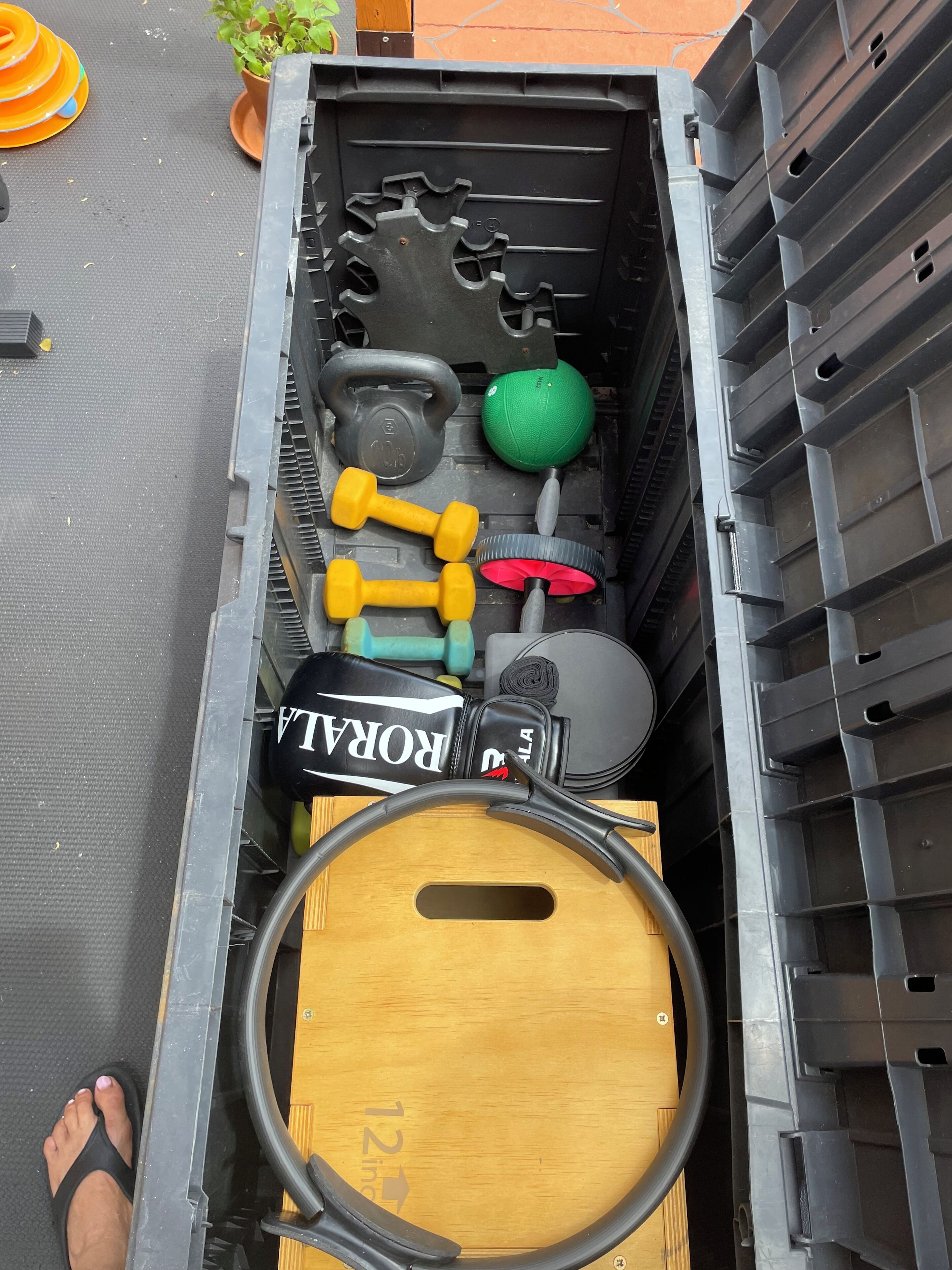 A storage container with various fitness equipment, including kettlebells, dumbbells, a medicine ball, and a yoga mat.