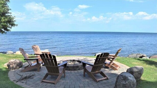 A circular outdoor seating area with Adirondack chairs and a fire pit, overlooking a body of water. - Newly renovated lakefront townhome with incredible views of Mille Lacs Lake (Onamia)