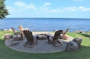 Un espace extérieur circulaire aménagé avec des chaises Adirondack et un foyer, offrant une vue sur une étendue d’eau.