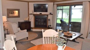 A living room with a fireplace, a television mounted above it, and a large window offering a view of the outdoors. - Newly renovated lakefront townhome with incredible views of Mille Lacs Lake (Onamia)