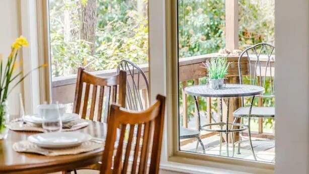 A dining area with a table set for two, a window with a view of greenery, and a small round table with chairs outside. - The Owl House cheerful, bright, updated 2B A-frame (Hot Springs)
