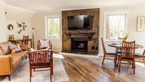 A living room with a stone fireplace, a television mounted above it, a leather sofa, a dining table with chairs, and a large window providing natural light. - The Owl House cheerful, bright, updated 2B A-frame (Hot Springs)