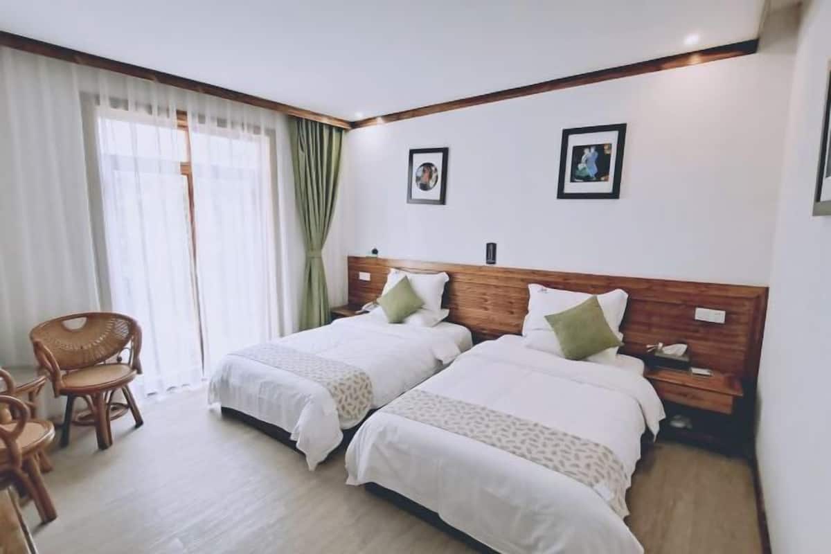 Une chambre d’hôtel avec deux lits, une tête de lit en bois, une chaise et une grande fenêtre avec des rideaux.