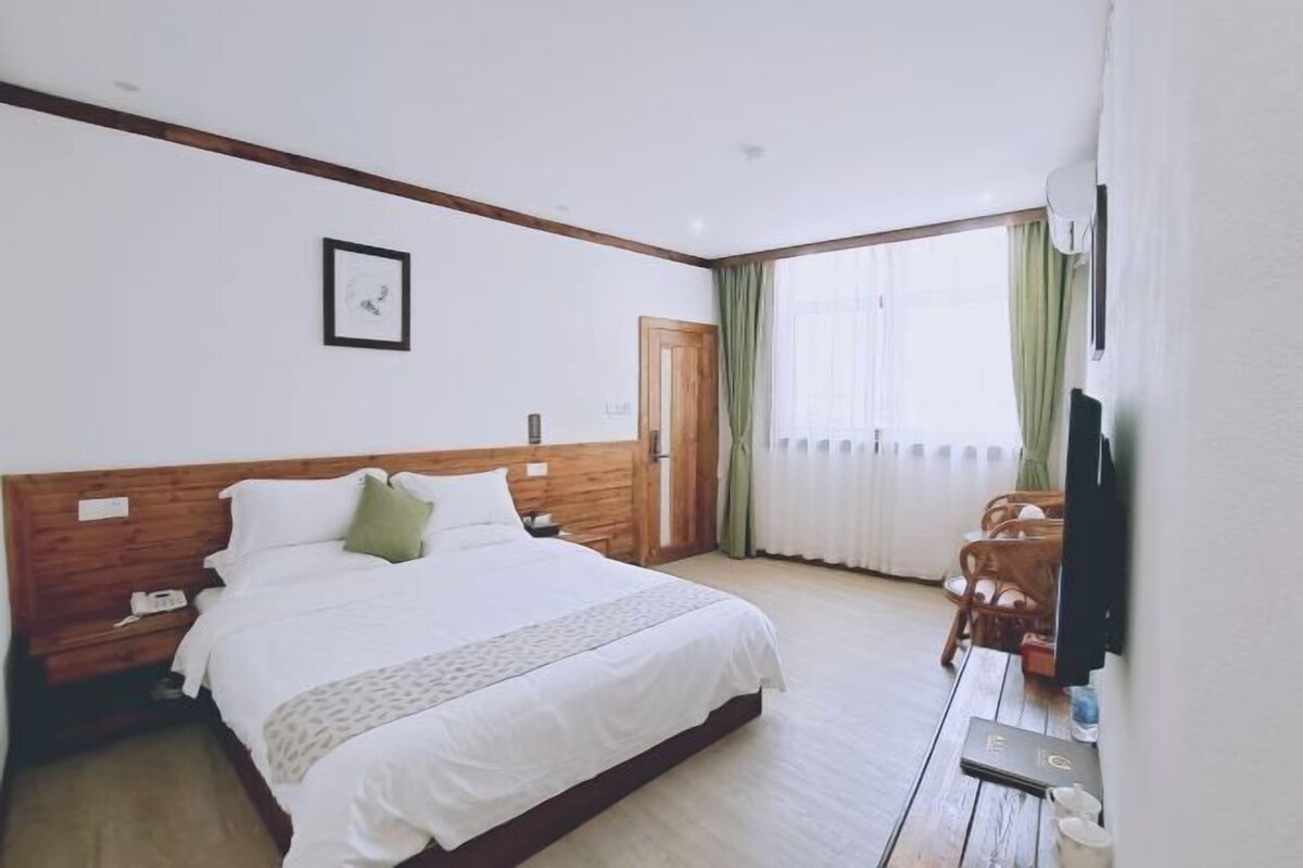 Een hotelkamer met een houten hoofdeinde, een bed met witte lakens, een klein nachtkastje, een tv en een raam met gordijnen.