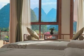 Een slaapkamer met een groot raam met uitzicht op de bergen, een bed met witte lakens en een klein tafeltje met een bloempot.