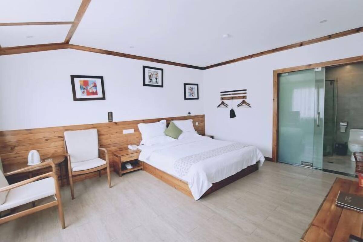 Une chambre moderne avec une tête de lit en bois, un lit avec une literie blanche, une chaise en bois, une table de chevet et une porte en verre donnant sur une salle de bain.