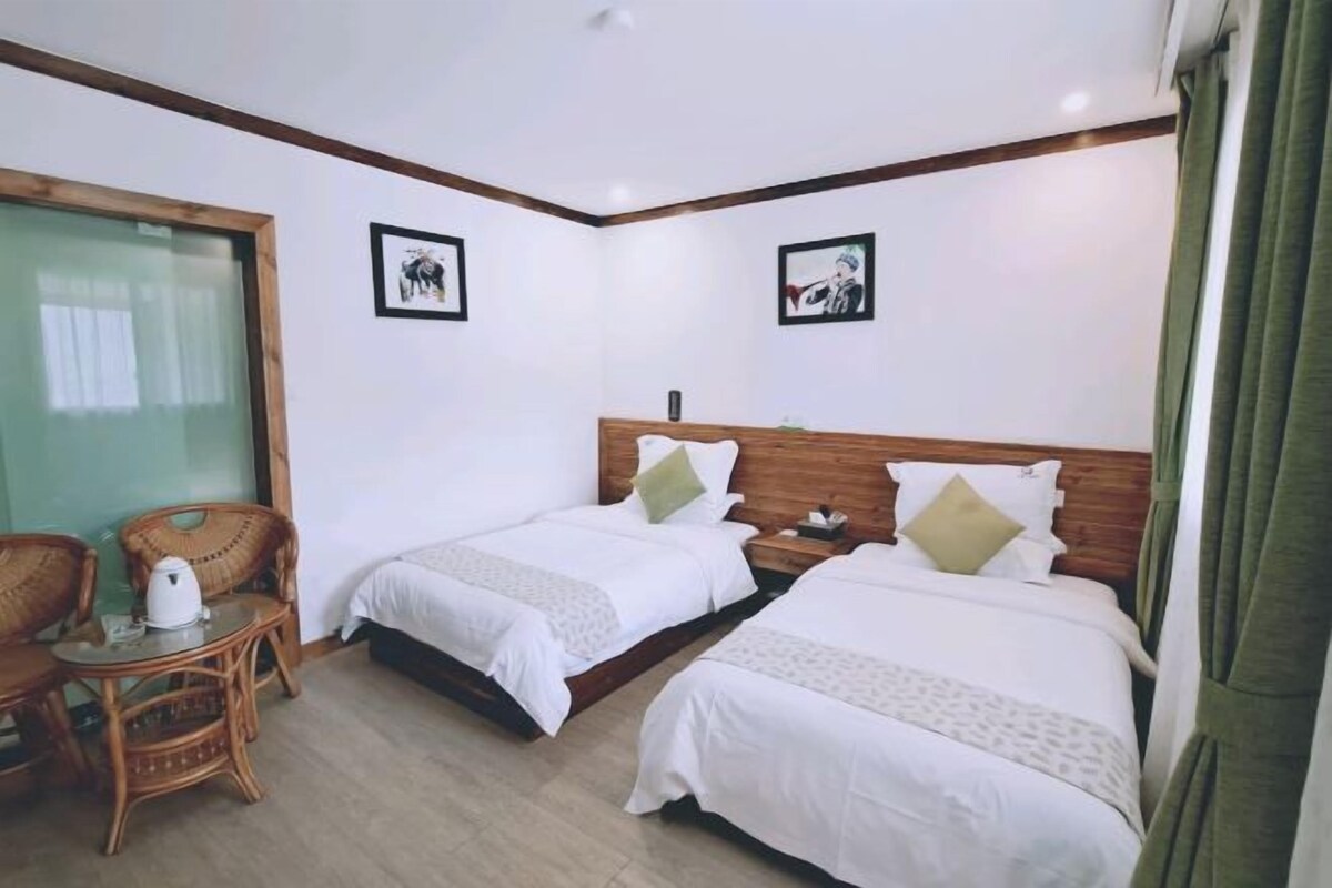 A hotel room with two single beds, a wooden headboard, a small table with a kettle, and two wicker chairs.