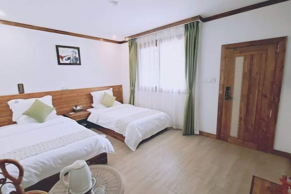 Une chambre d’hôtel avec deux lits, une tête de lit en bois, une fenêtre avec des rideaux, une porte et une petite table avec une bouilloire.