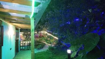 Un espacio al aire libre cubierto con un suelo de madera, rodeado de follaje e iluminado con luces azules.