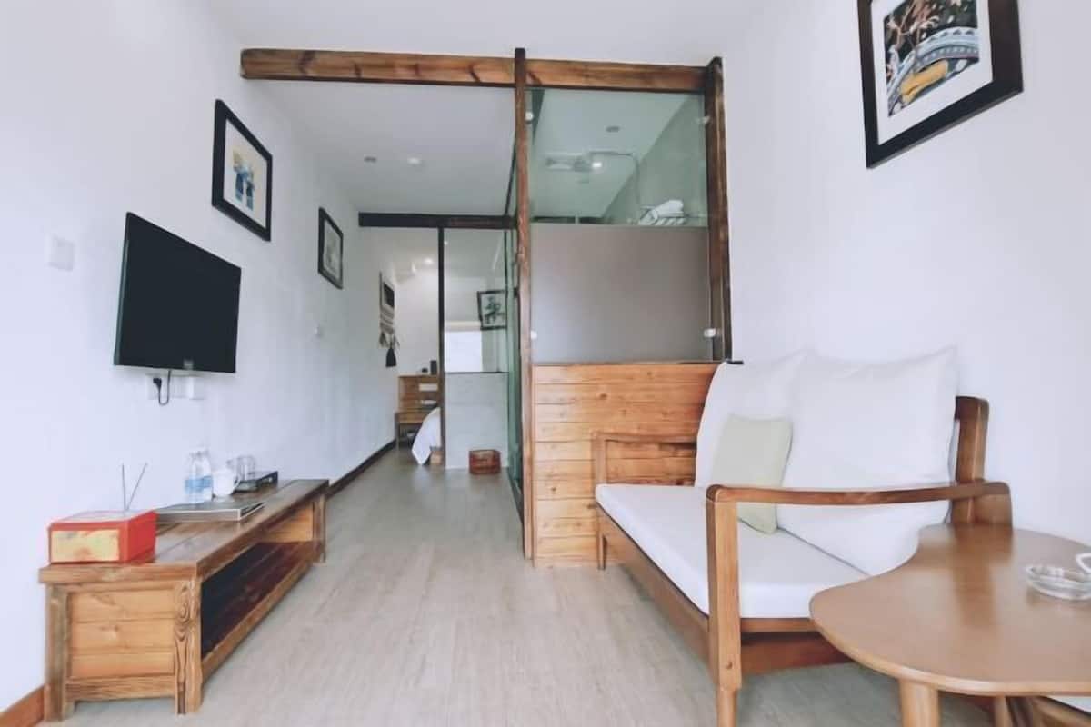 Une chambre d’hôtel moderne avec un canapé en bois, une petite table en bois, un téléviseur fixé au mur et une porte vitrée donnant sur un balcon.
