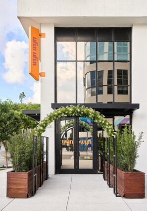 A modern building entrance with a wooden planter walkway, a clear sky, and a reflection of a building in the glass facade. - Arlo Wynwood Miami (Miami)