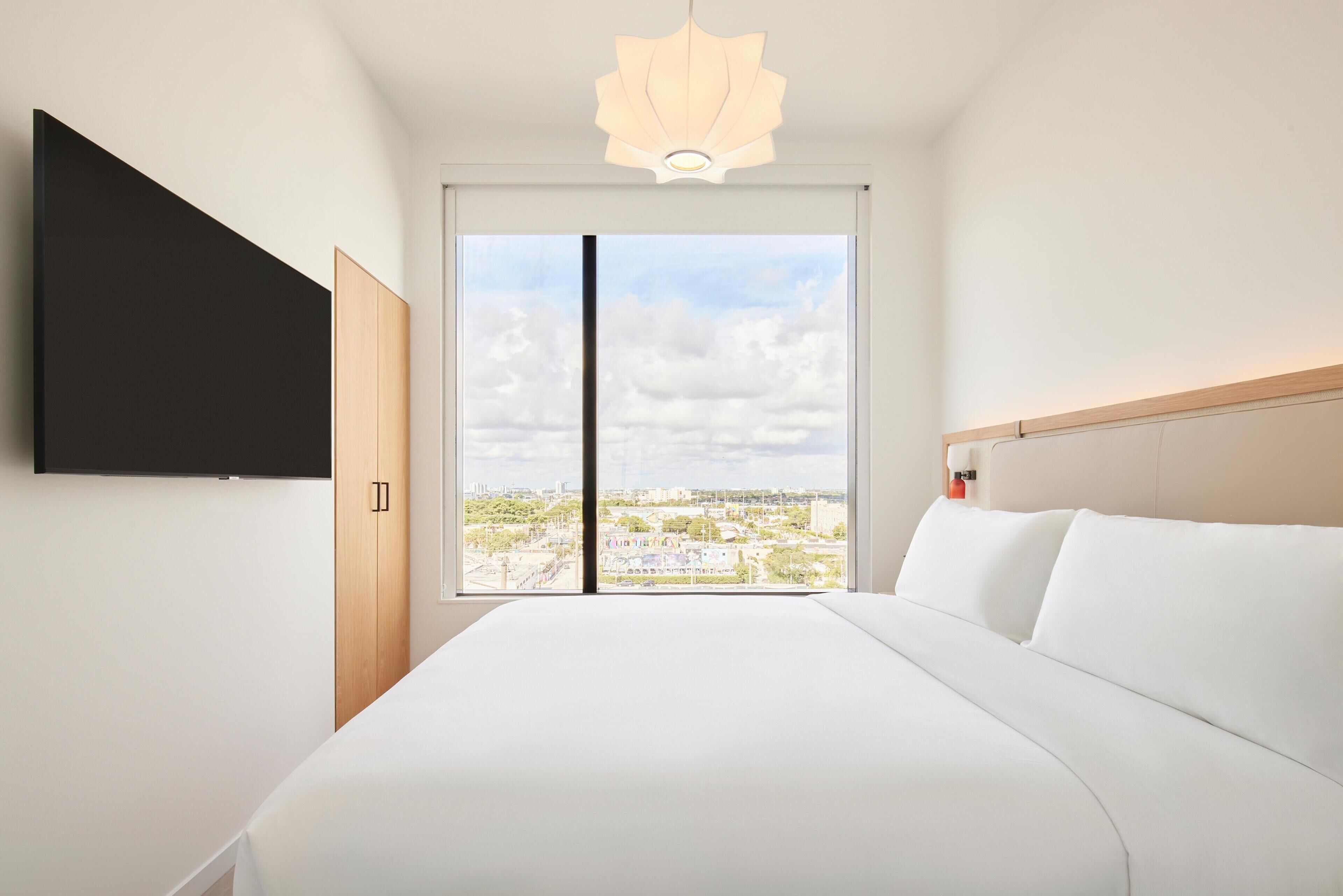 A modern hotel room with a large bed, a flat-screen TV mounted on the wall, and a city view through the window.