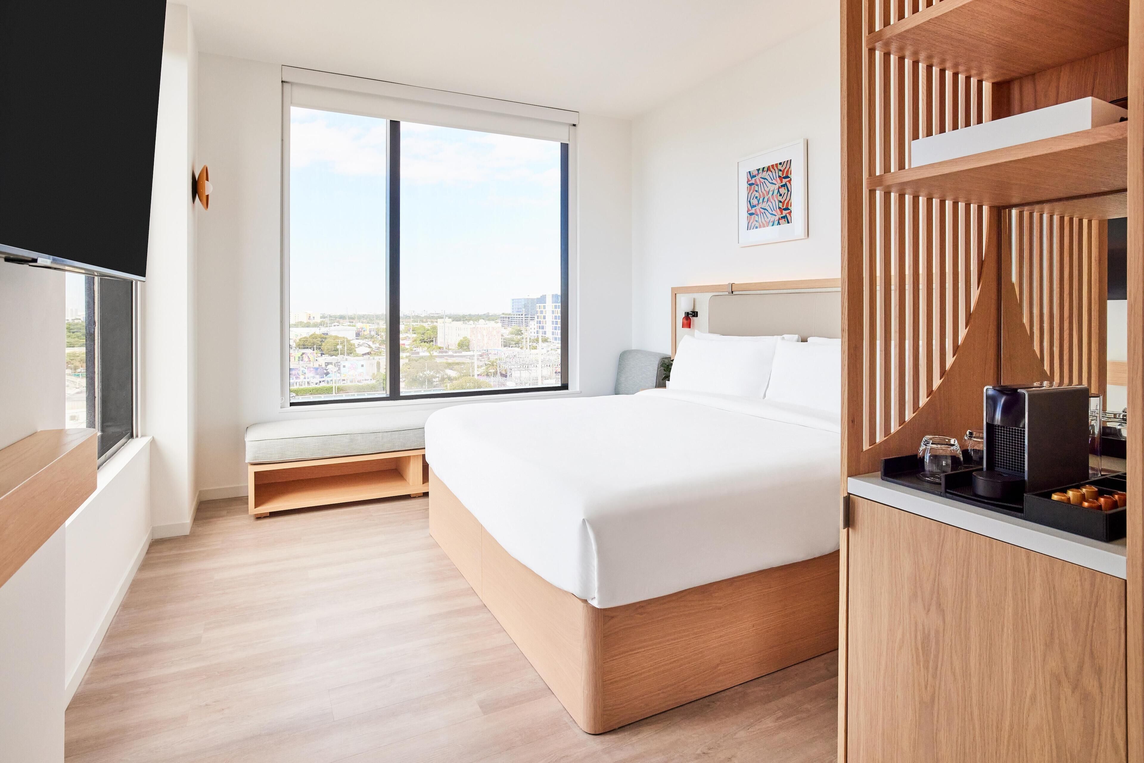 A hotel room with a large bed, a window with a city view, a small seating area, and a wooden cabinet with a coffee machine.