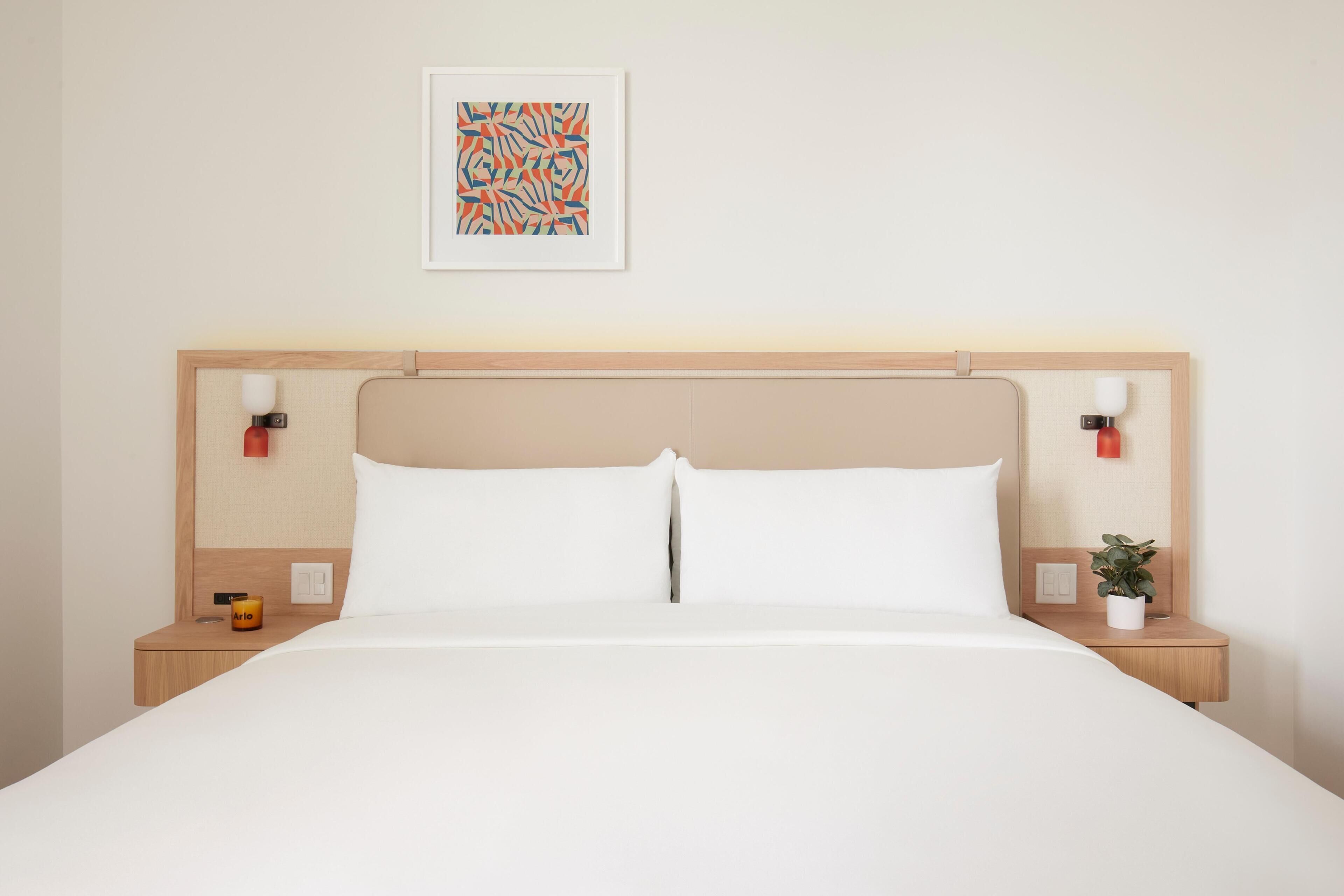 A hotel room with a bed, nightstands, a wall-mounted lamp, a potted plant, and a framed abstract art piece.