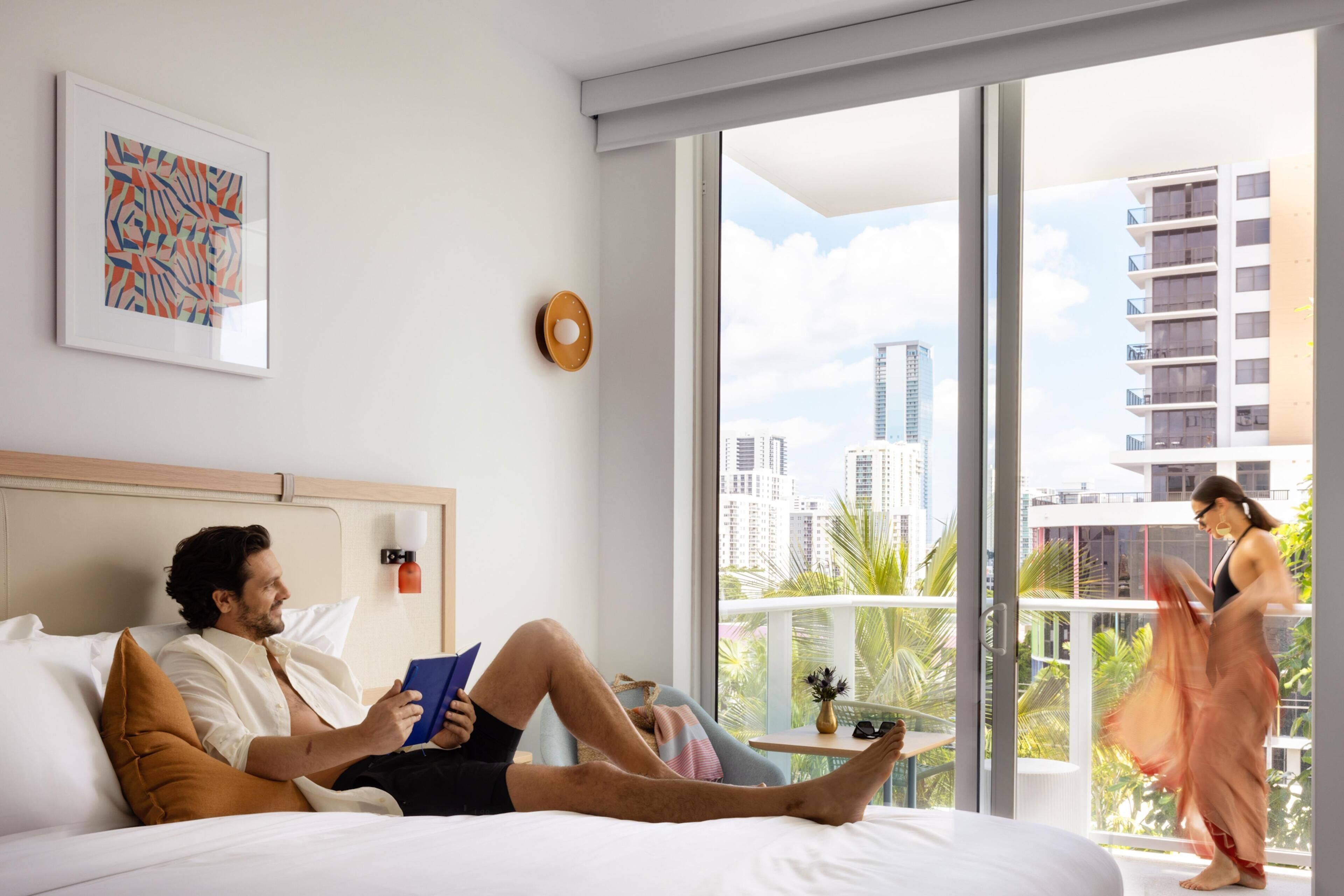 A hotel room with a bed, a reading lamp, a framed abstract painting, and a view of a cityscape through large windows.