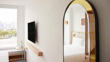 Quarto de hotel com cama, televisão fixada na parede, mesinha com uma planta e janela com vista para a cidade.
