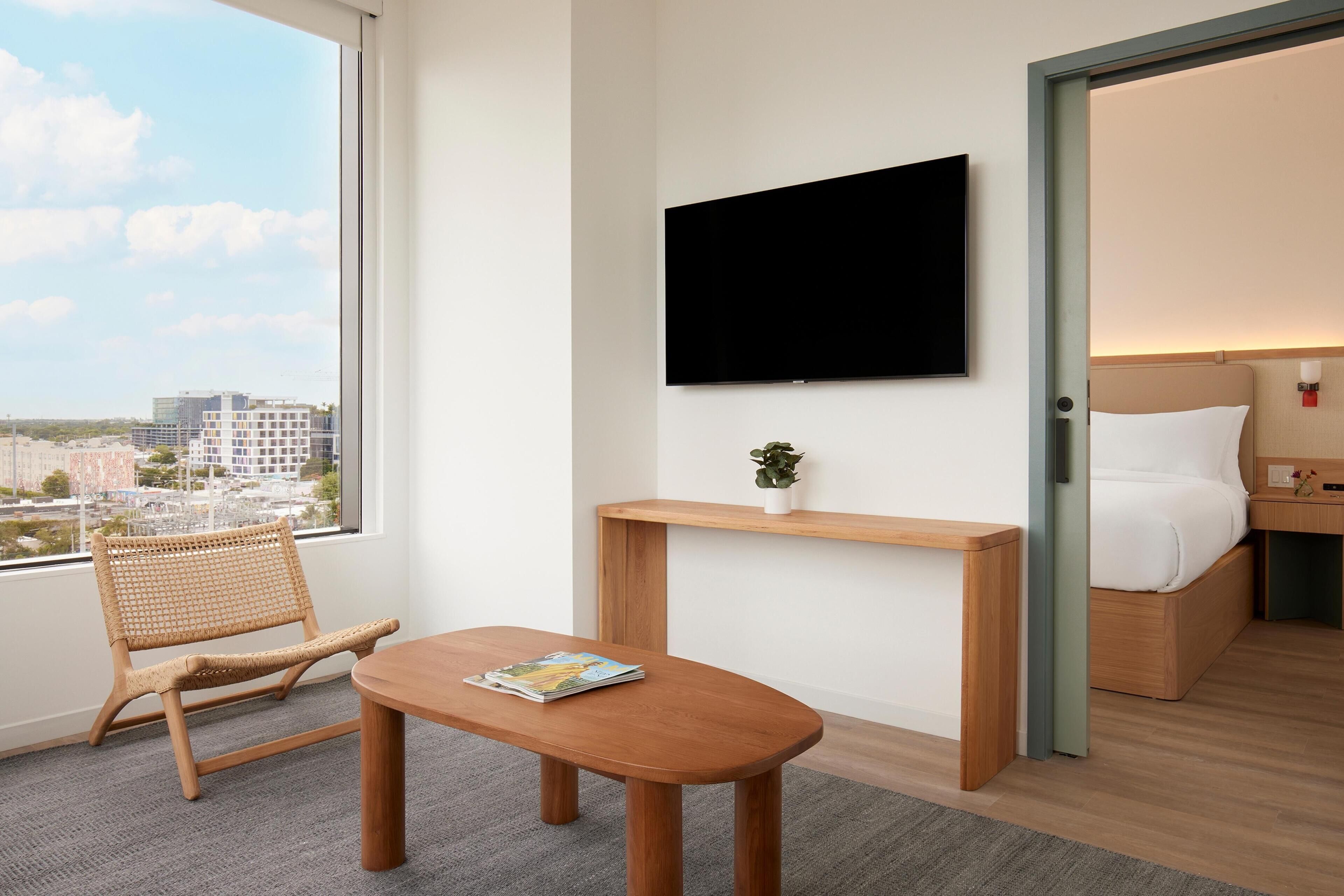 A hotel room with a large window, a flat-screen TV mounted on the wall, a wooden bedside table with a plant, a wicker chair, and a bed with white bedding.