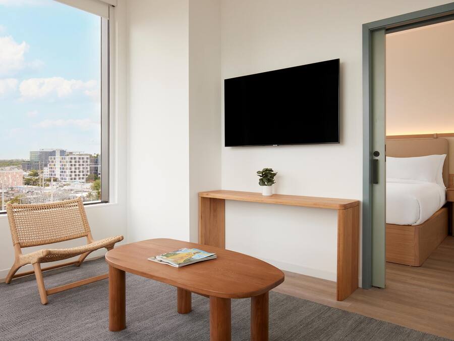 A hotel room with a large window, a flat-screen TV mounted on the wall, a wooden bedside table with a plant, a wicker chair, and a bed with white bedding.