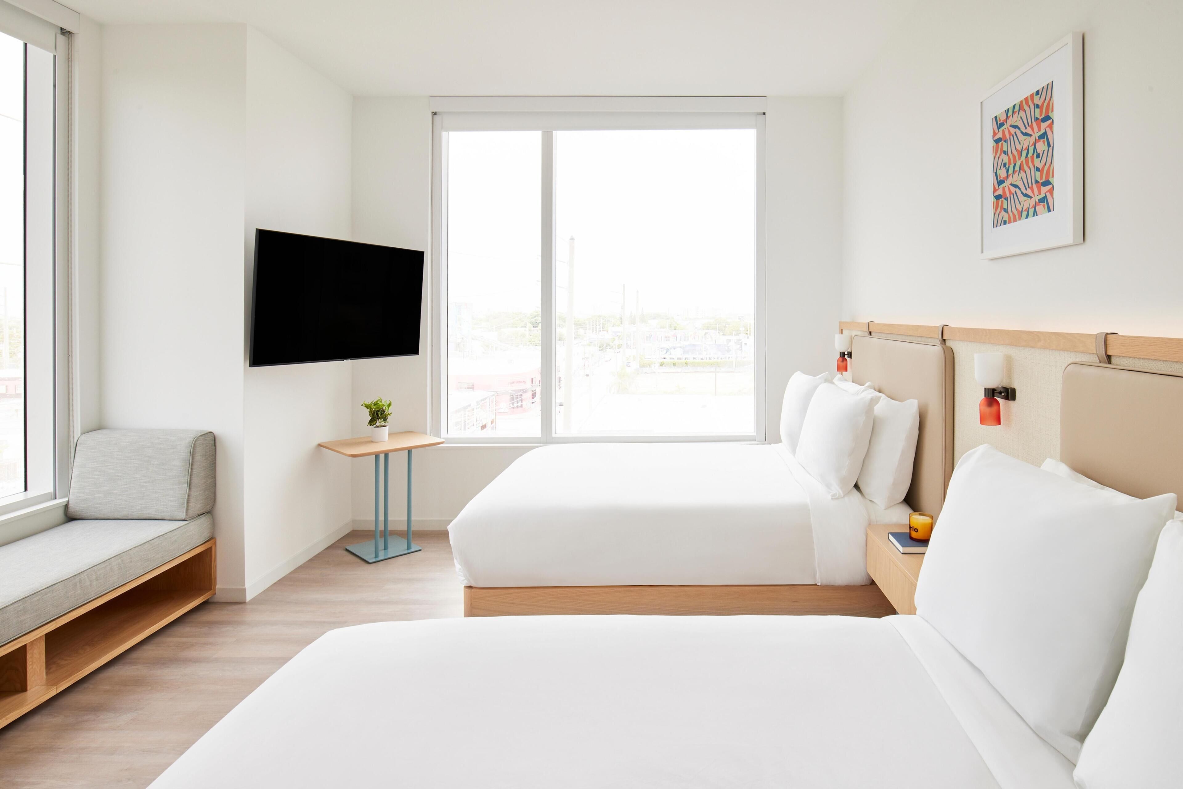 A hotel room with a bed, a seating area, a TV, and a window with a city view.