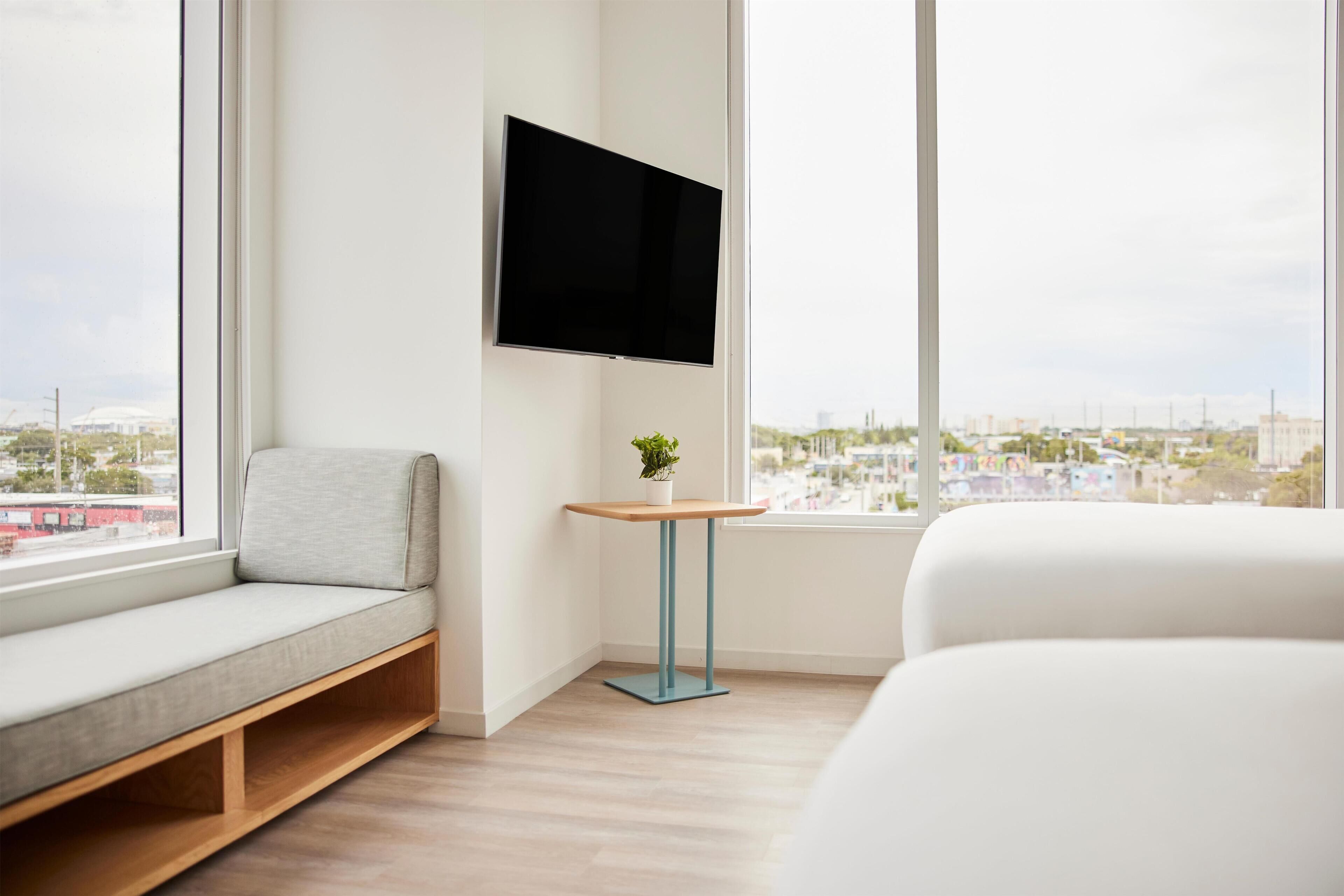 Ein modernes Hotelzimmer mit großem Fenster, einem Wand-Flachbildfernseher, einer Couch, einem kleinen Tisch mit Pflanze und einem Bett.