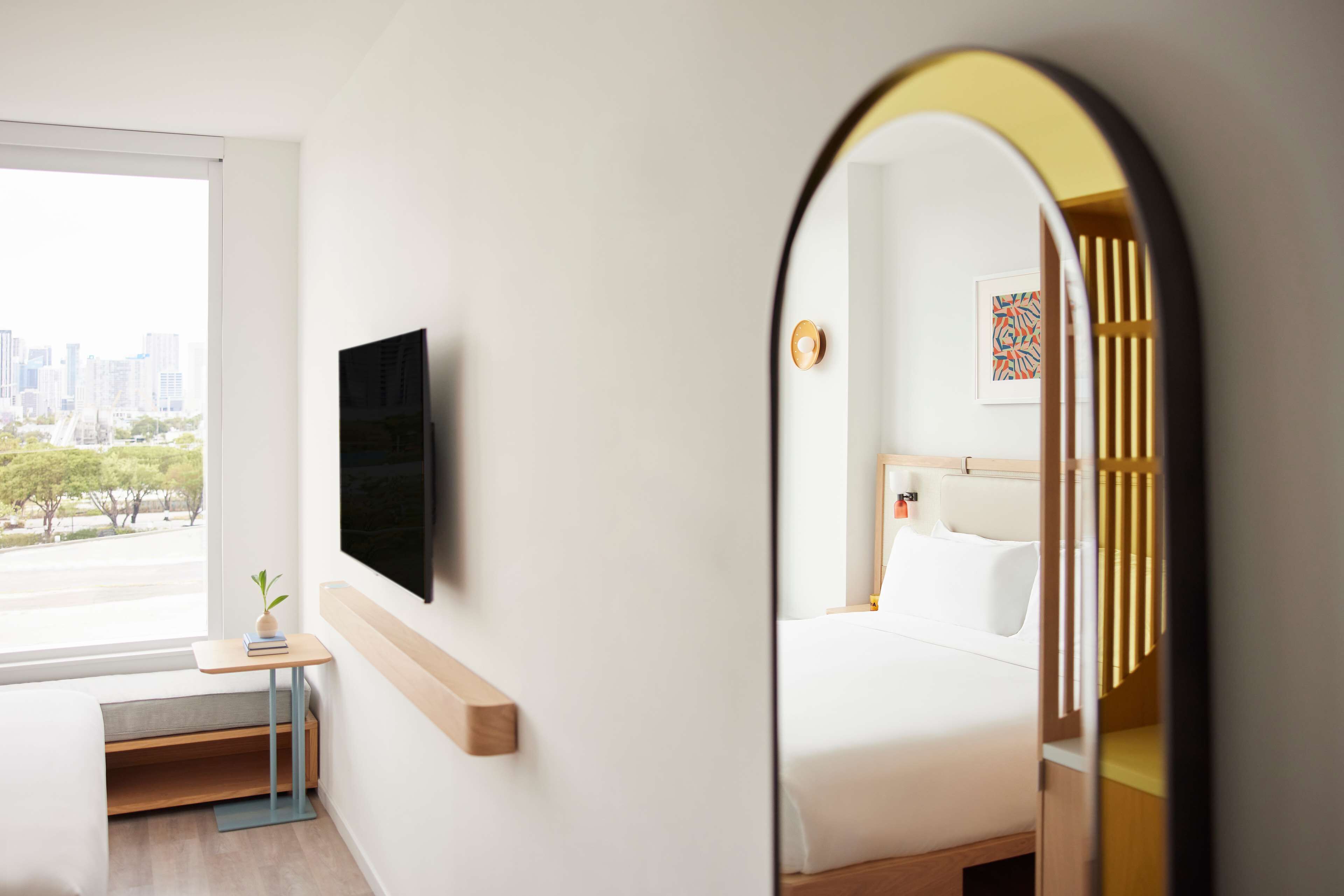 Een hotelkamer met een bed, een nachtkastje met een plant, een wandtelevisie en een grote spiegel die de kamer reflecteert.