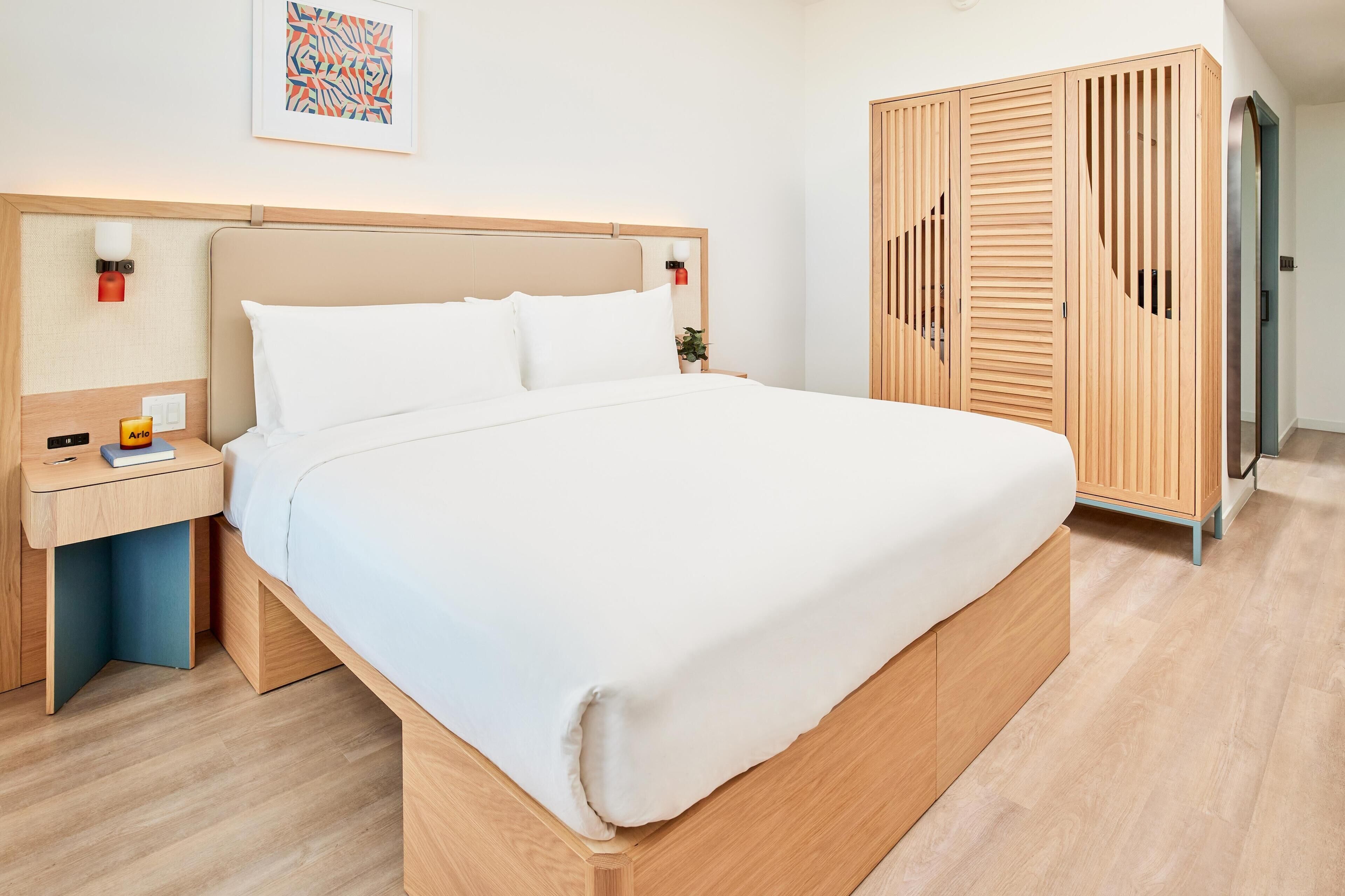 Une chambre d’hôtel moderne avec un grand lit, une armoire en bois, une table de chevet avec une lampe et une petite plante en pot.