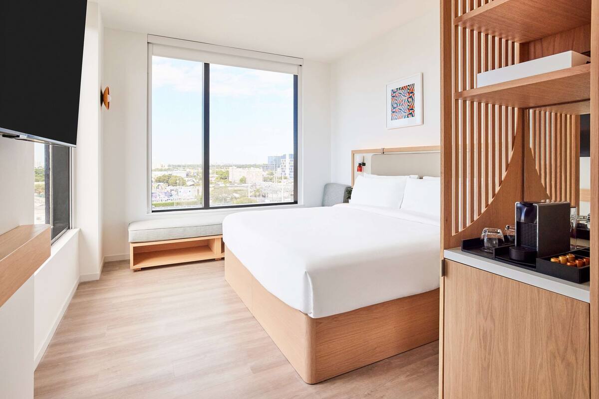 A modern hotel room with a large bed, a window with a city view, a wooden desk with a coffee maker, and a small seating area.